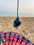 Handcrafted Blue Quartz spiral pendulum  suspended above a pebble beach, wrapped in titanium wire on a soft grey cord with the sea in the background.