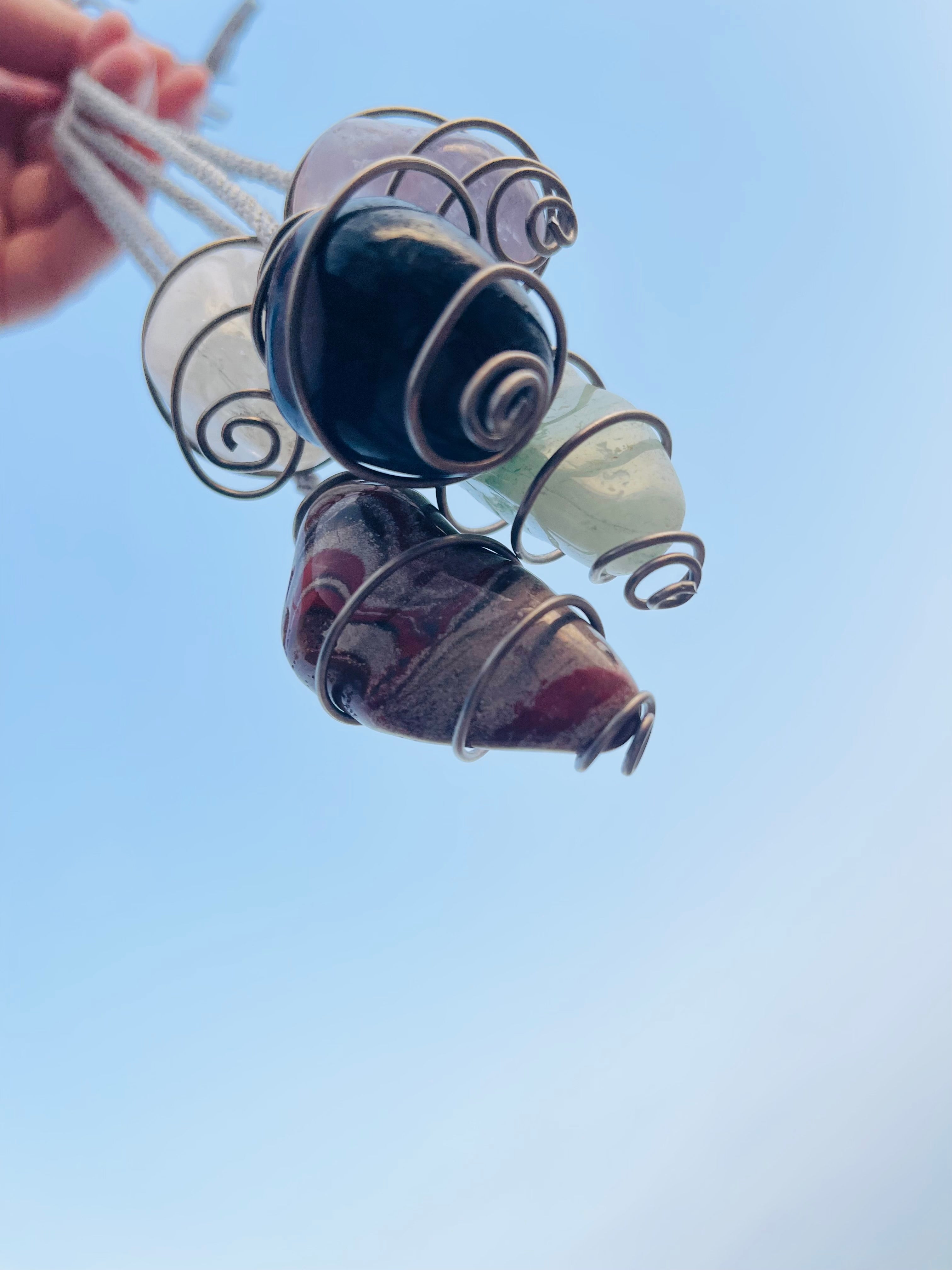 Blue Quartz spiral pendulum wrapped in titanium wire, shown alongside other handcrafted crystal pendulum necklaces against a bright blue sky.