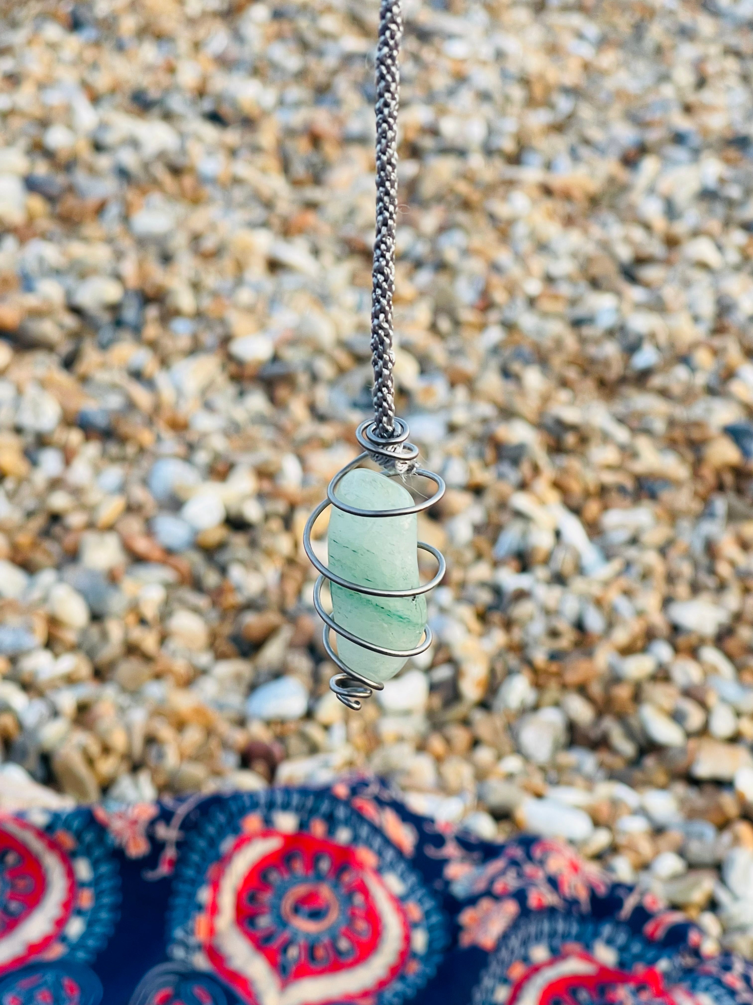 Green Aventurine crystal pendulum handcrafted with Grade 1 titanium wire hanging above a pebble beach on the Kent coast.