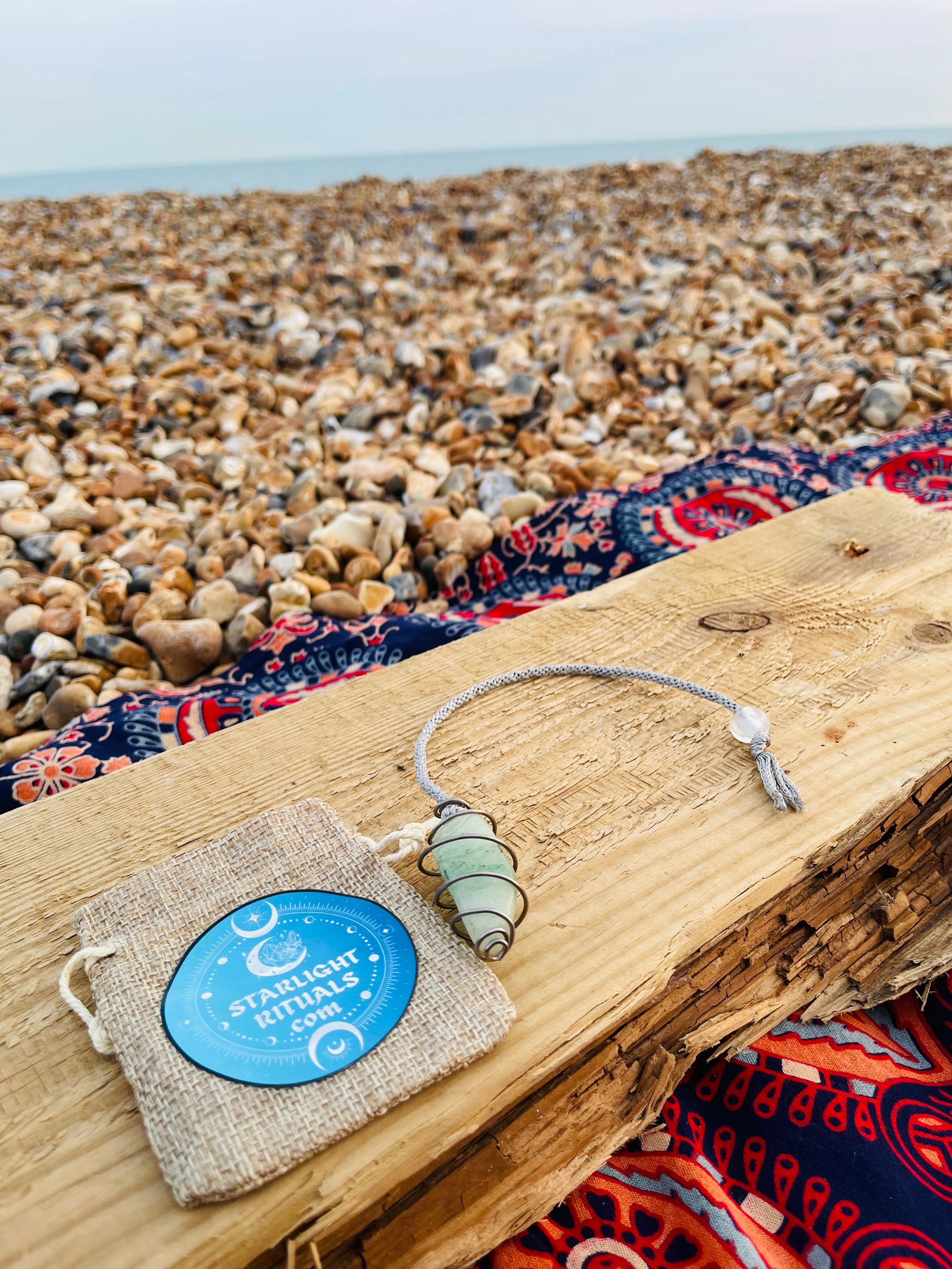 Green Aventurine crystal pendulum with a hand-braided rayon cord displayed beside a Starlight Rituals linen gift pouch on driftwood at a pebble beach.