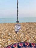 Handcrafted Light Amethyst crystal pendulum wrapped in Grade 1 titanium wire, hanging above a pebble beach on the Kent coast at sunset.