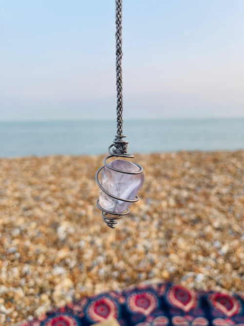 Handcrafted Light Amethyst crystal pendulum wrapped in Grade 1 titanium wire, hanging above a pebble beach on the Kent coast at sunset.