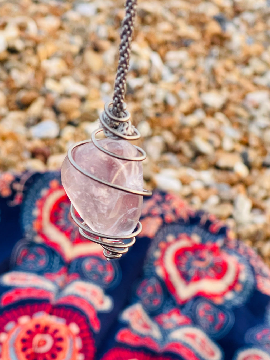 Close-up of a handcrafted Light Amethyst crystal pendulum wrapped in Grade 1 titanium wire, suspended above a pebble beach with the Starlight Rituals patterned blanket below.