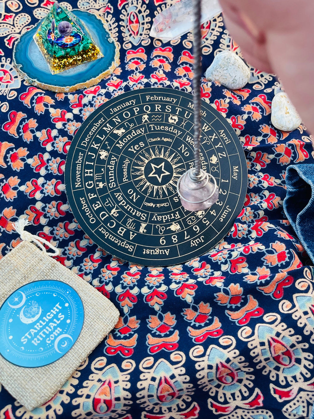 Handcrafted Light Amethyst crystal pendulum suspended above a dowsing board during a pendulum reading, with the Starlight Rituals linen gift pouch displayed alongside.