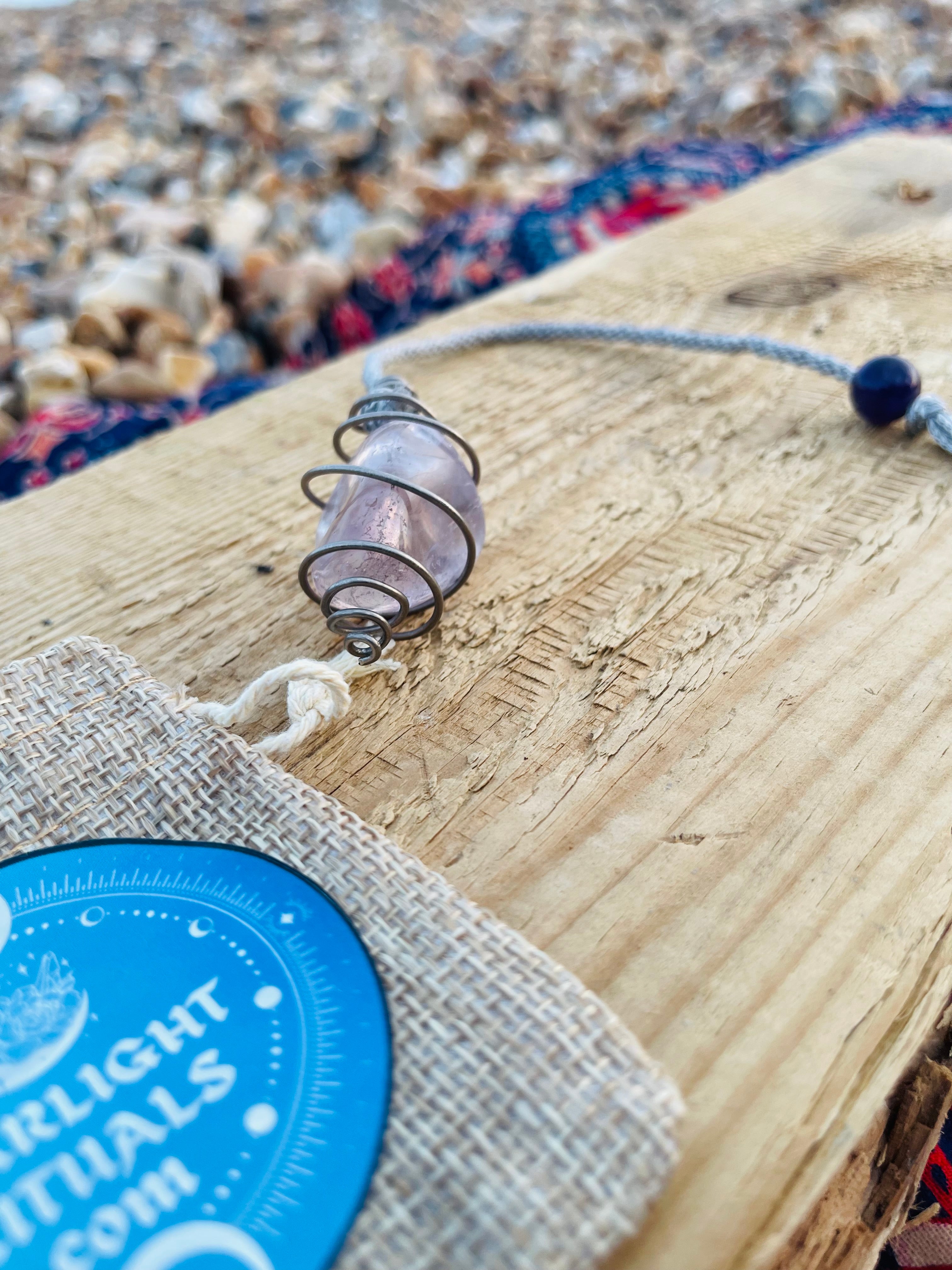 Handcrafted Light Amethyst crystal pendulum wrapped in Grade 1 titanium wire, resting on driftwood with its hand braided rayon cord and Starlight Rituals linen gift pouch.
