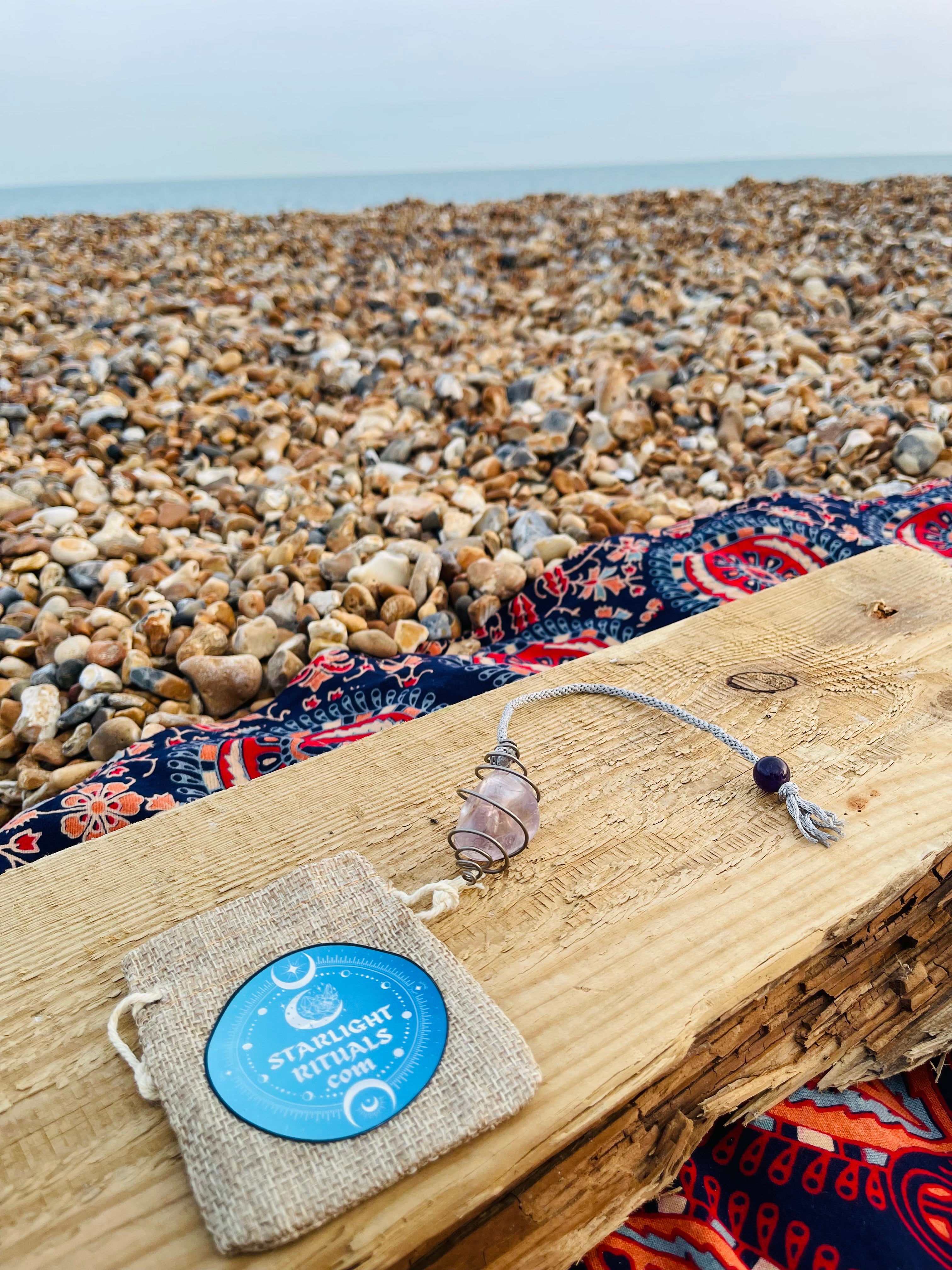Handcrafted Light Amethyst crystal pendulum with hand braided rayon cord, presented alongside a Starlight Rituals linen gift pouch on driftwood overlooking the Kent coast.