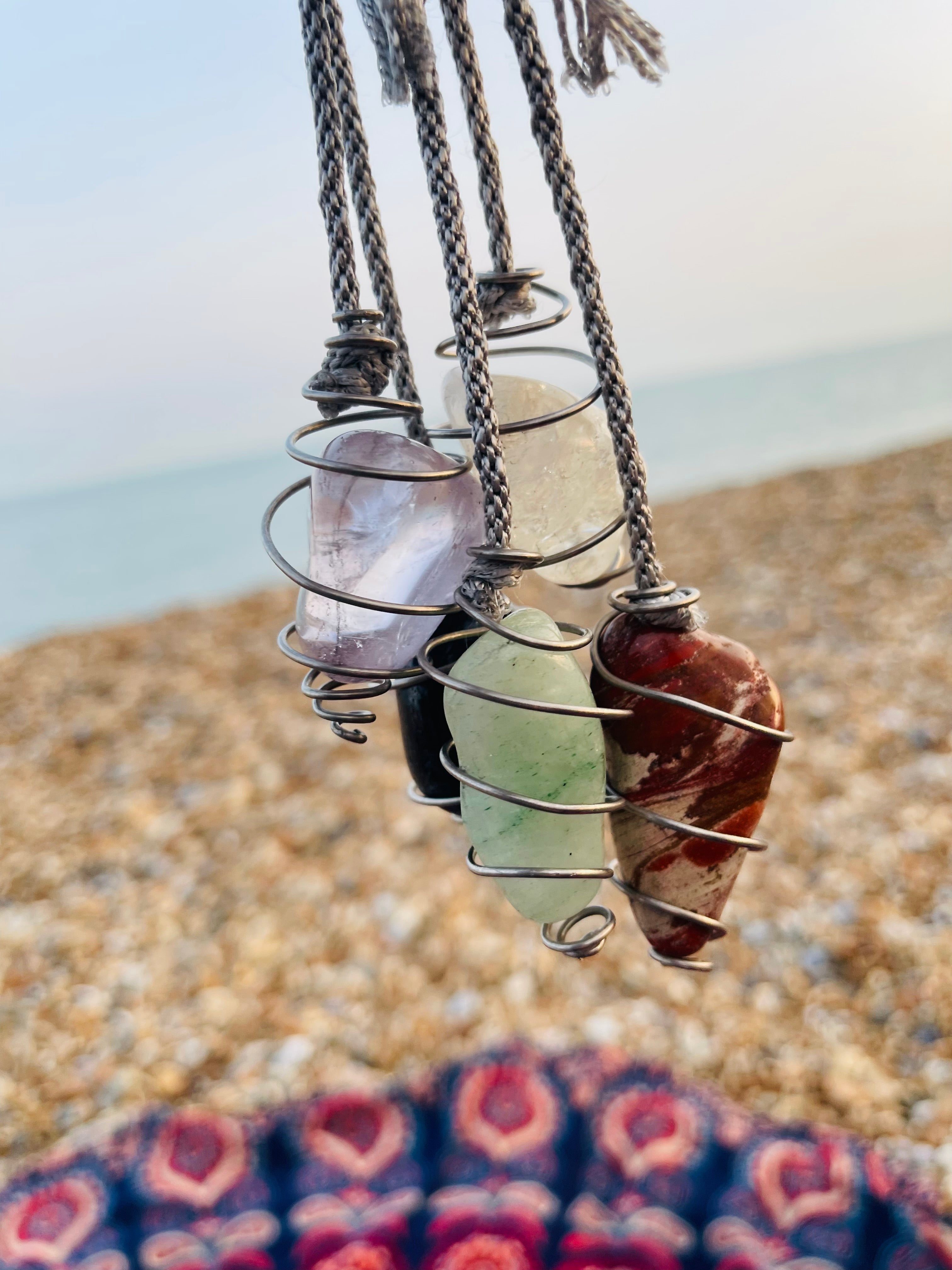 Light Amethyst crystal pendulum displayed alongside other handcrafted Starlight Rituals crystal pendulums, each wrapped in Grade 1 titanium wire, above a pebble beach on the Kent coast.