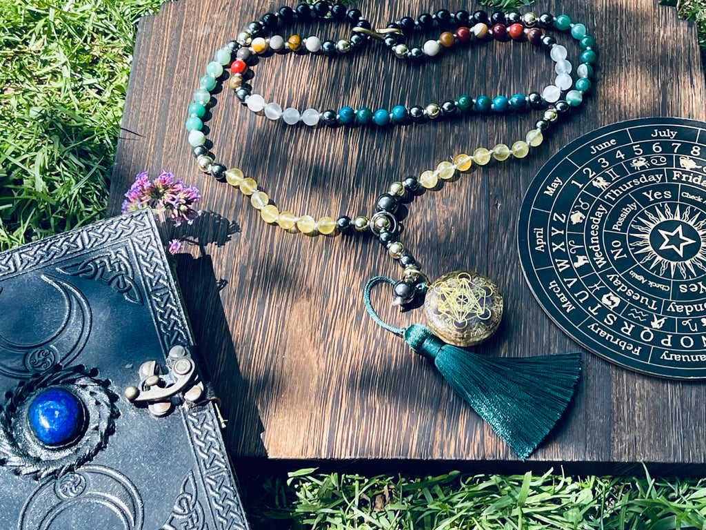 Path of Prosperity crystal mala displayed with a journal and ritual wheel, featuring a Metatron's Cube talisman and emerald green tassel.