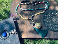 Path of Prosperity crystal mala displayed with a journal and ritual wheel, featuring a Metatron's Cube talisman and emerald green tassel.