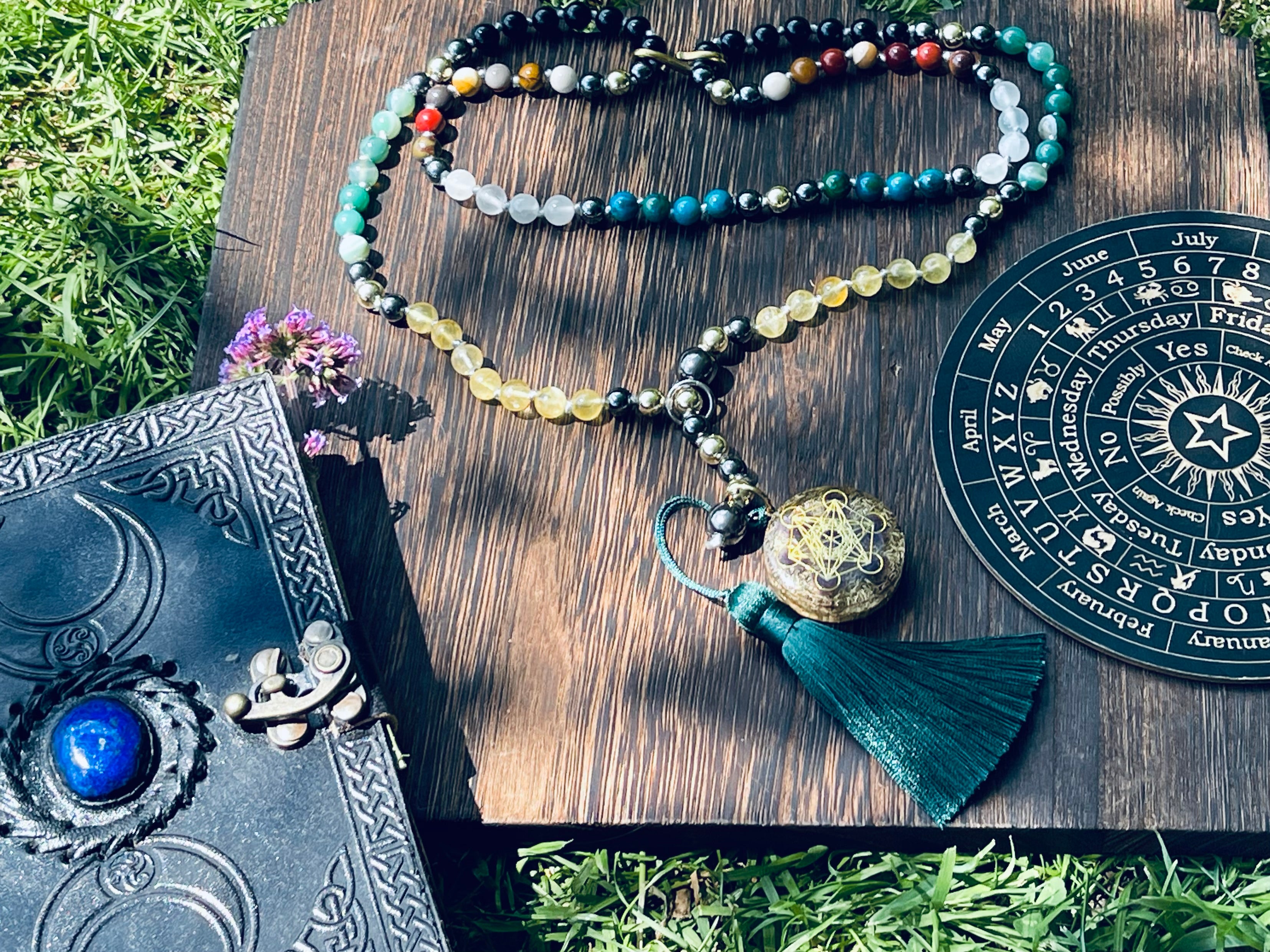 Path of Prosperity crystal mala displayed with a journal and ritual wheel, featuring a Metatron's Cube talisman and emerald green tassel.