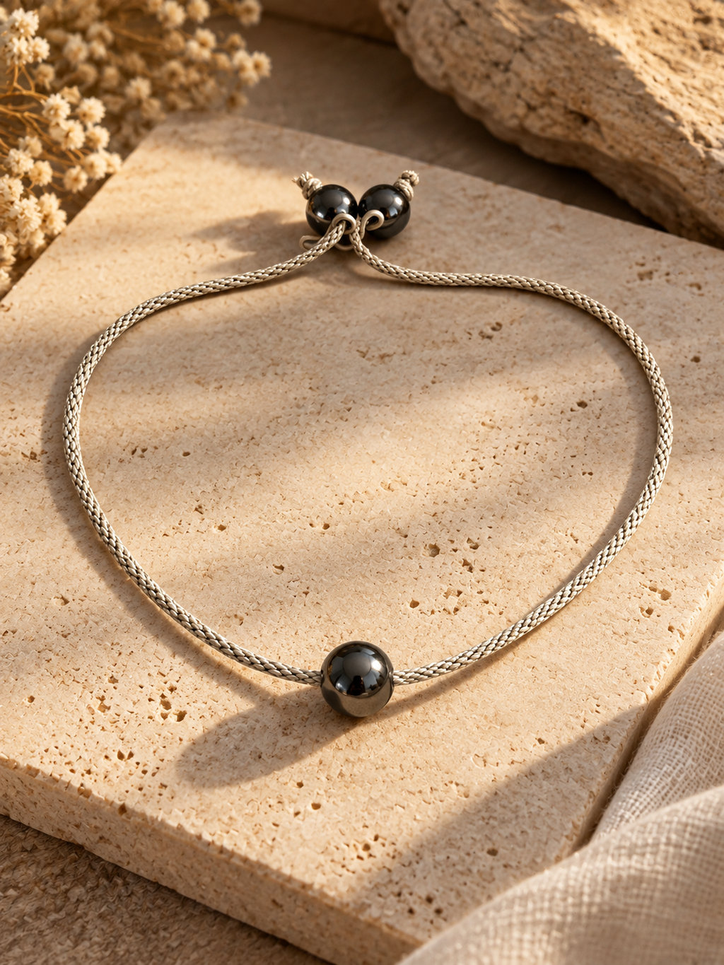 Handcrafted adjustable bracelet by Starlight Rituals featuring a polished hematine bead on a silver hand-braided cord, with two hematine beads at the sliding closure, displayed on natural beige stone.