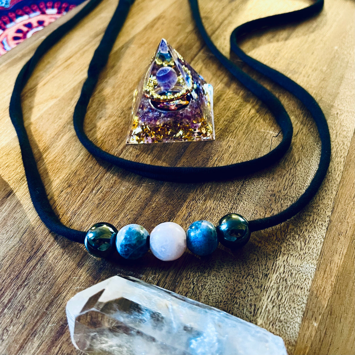 Handmade Crystal Comfort Belt featuring five hematine, sodalite and rose quartz beads on soft black bamboo jersey, displayed on warm wood with an amethyst orgonite pyramid and clear quartz crystal.