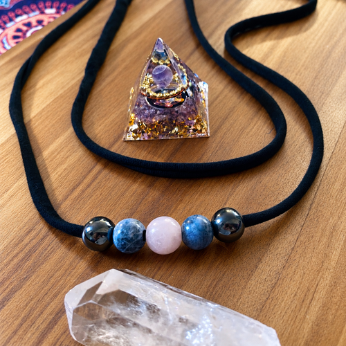 Handmade Crystal Comfort Belt featuring five hematine, sodalite and rose quartz beads on soft black bamboo jersey, displayed on warm wood with an amethyst orgonite pyramid and clear quartz crystal.