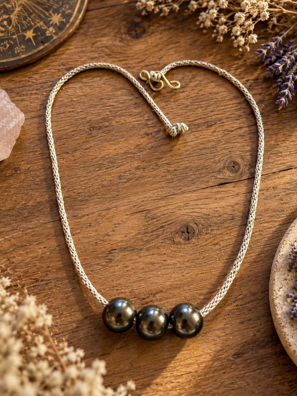 Handcrafted Starlight Rituals necklace with three polished hematine beads on a silver-toned braided cord, arranged in a heart shape on rustic wood with dried flowers and crystals.